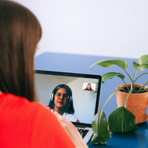 Woman in red shirt video chatting on laptop with plant nearby at home.